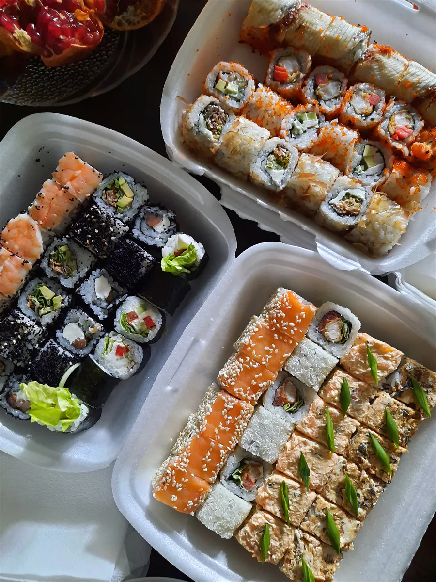 Three takeout containers filled with a variety of sushi rolls, including salmon nigiri, sesame-topped rolls, and assorted maki, with a pomegranate in the background.